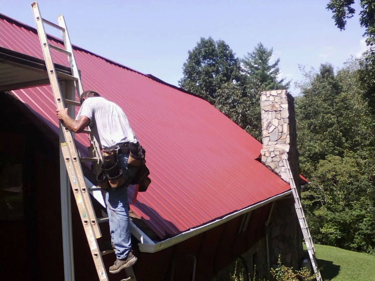 Roofer on a ladder beside a red metal roof and stone chimney — Soffit & Fascia Repair by Roofing Brothers in Marble Rock